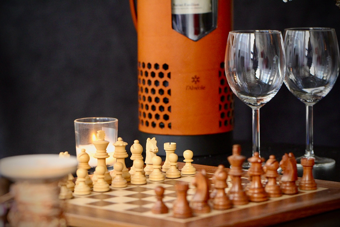 Un verre à vin et une bouteille de vin sur une table, avec L'Alvéole en arrière-plan et un jeu d'échec.